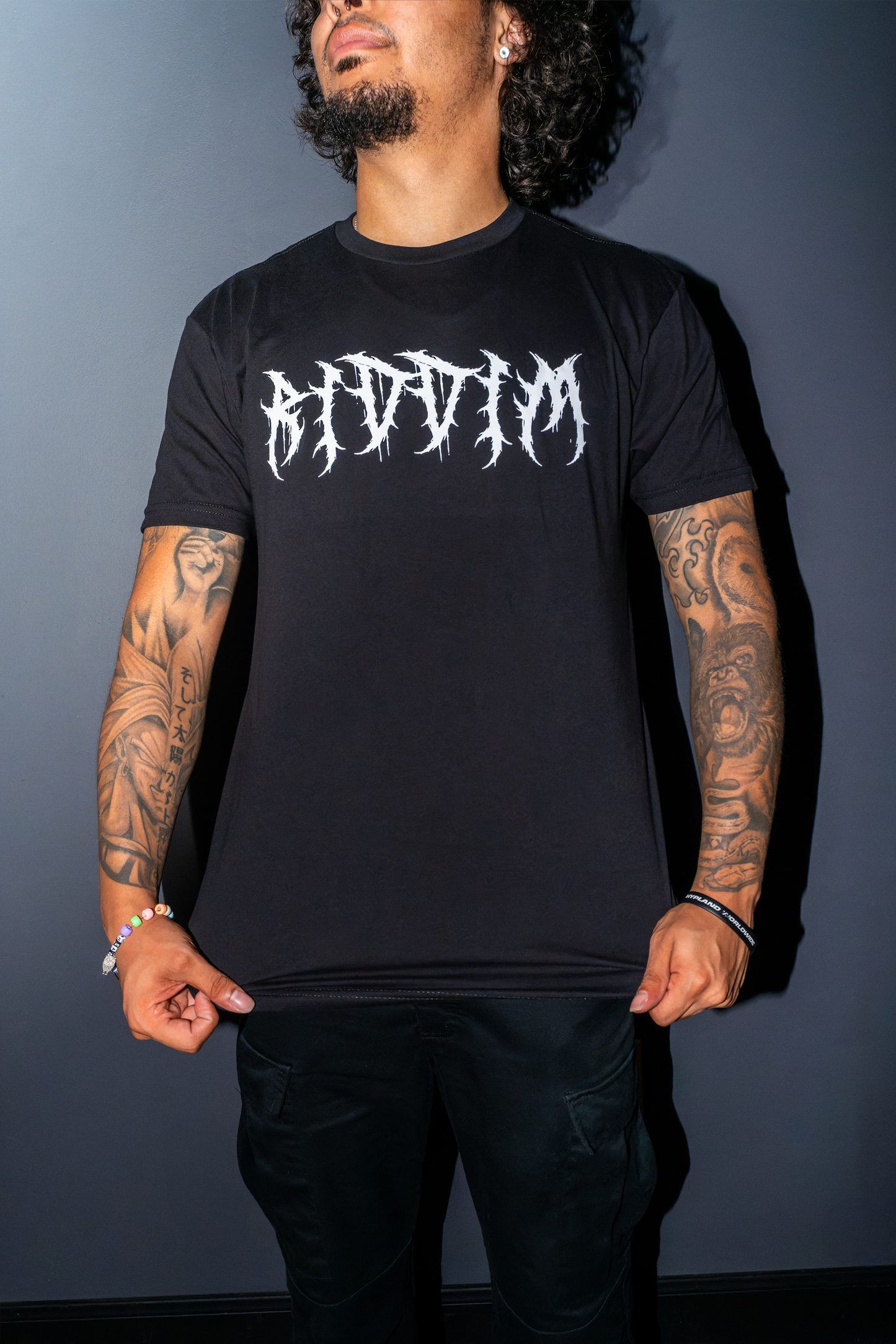 A man wearing a black Riddim Fallen Ange lt-shirt with a barbed white gothic-style 'Riddim' graphic on the chest, posing against a dark gray wall.