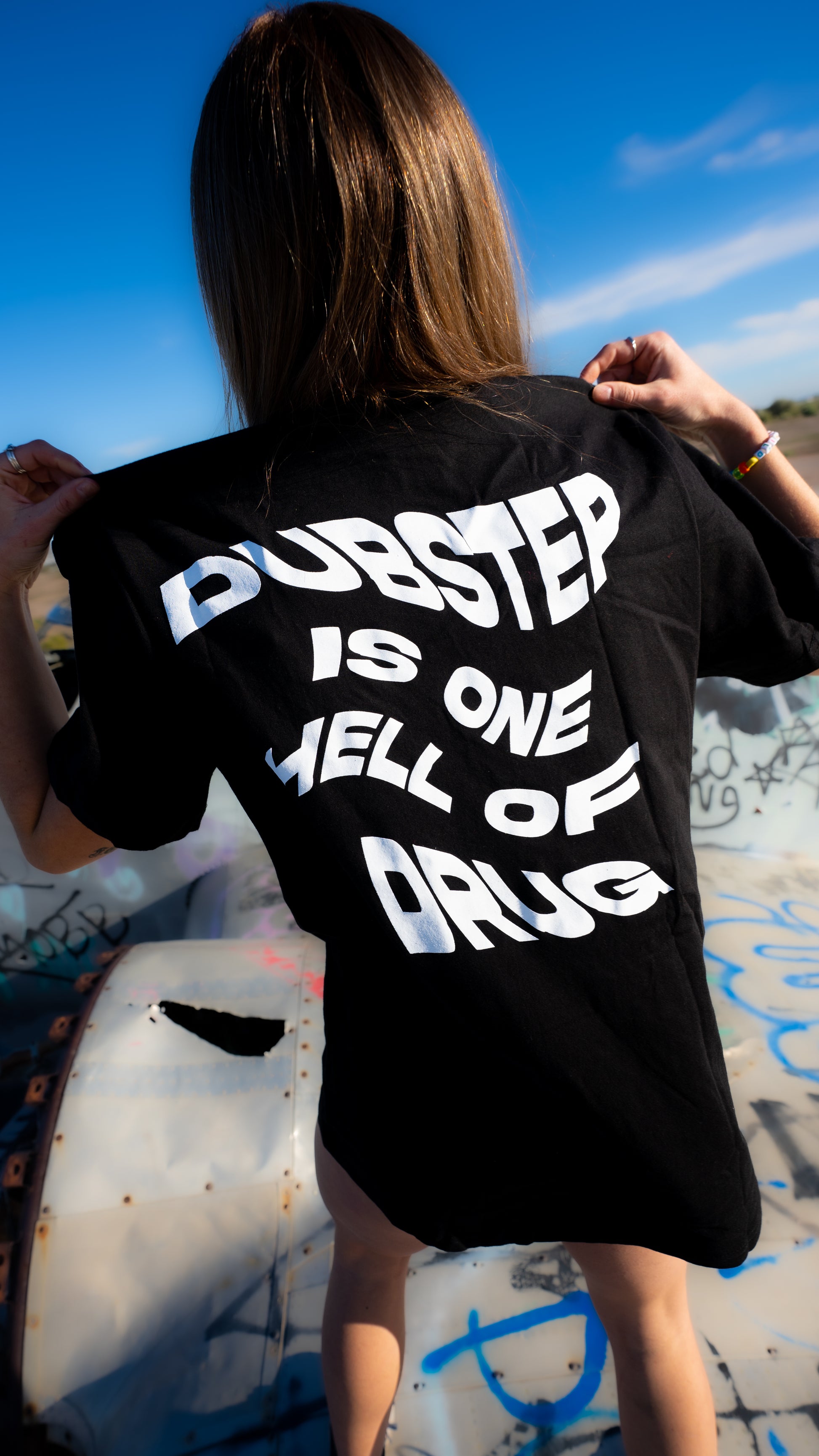 A person viewed from behind wearing an oversized black t-shirt with bold white text that reads 'DUBSTEP IS ONE WELL OF DRUG'. The background is an outdoor scene with graffiti and a blue sky.