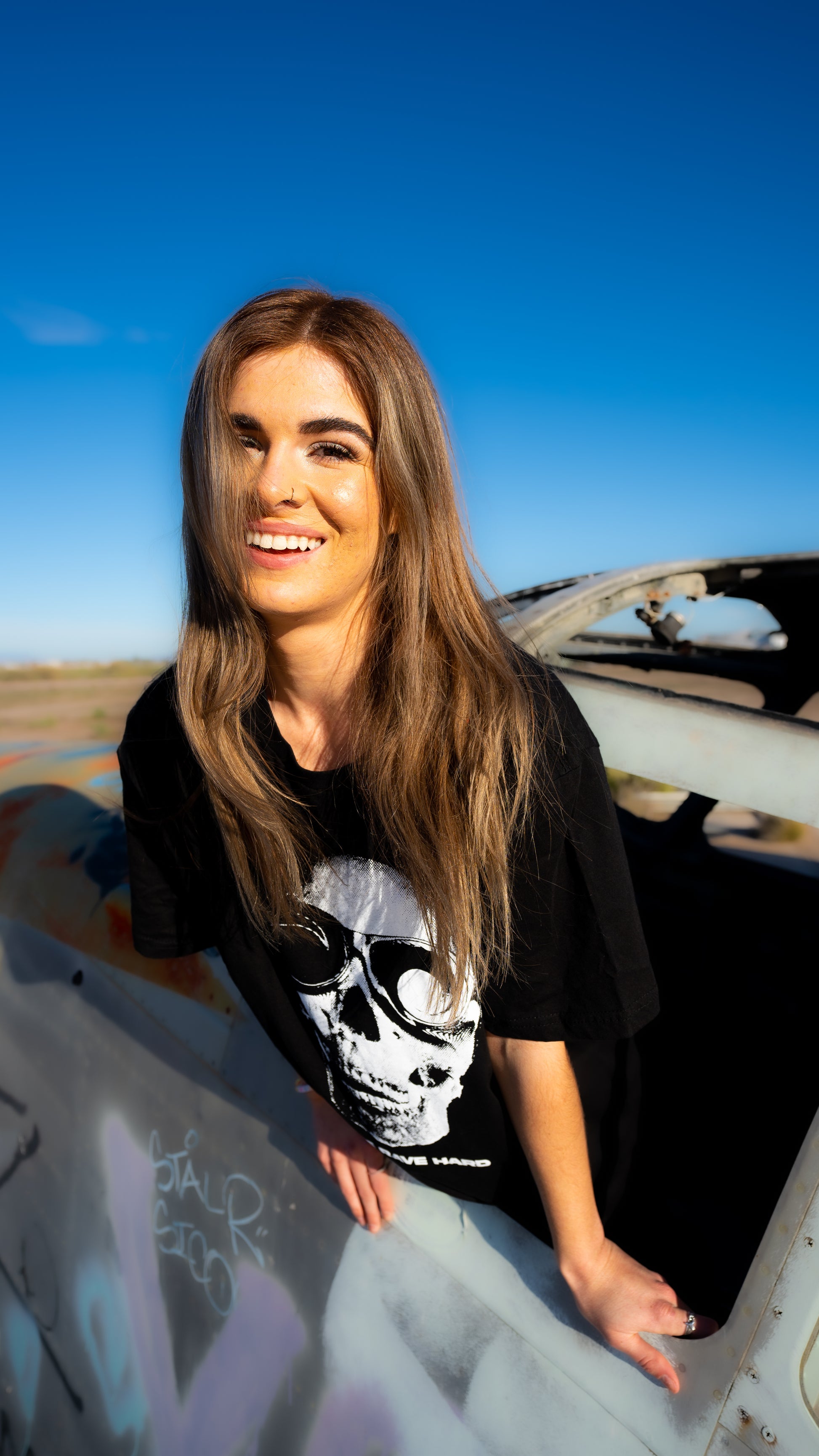 A woman with long brown hair leans against the open door of a graffiti-sprayed car outdoors under a clear blue sky, wearing a black oversized t-shirt with a white skull wearing sunglasses graphic.