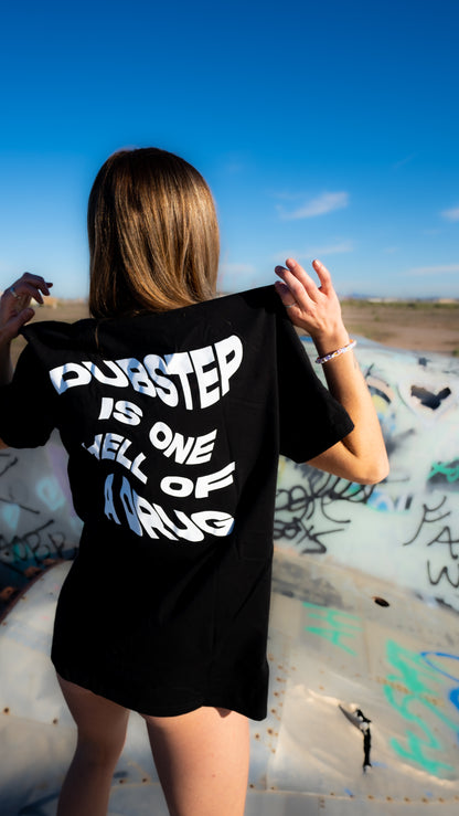A woman stands outdoors in a desert-like open area holding up a black oversized T-shirt from the back, which features the text 'DURSTEP IS ONE HELL OF A DRUG' in bold white lettering. The background shows a graffiti-covered metal surface under a clear blue sky.