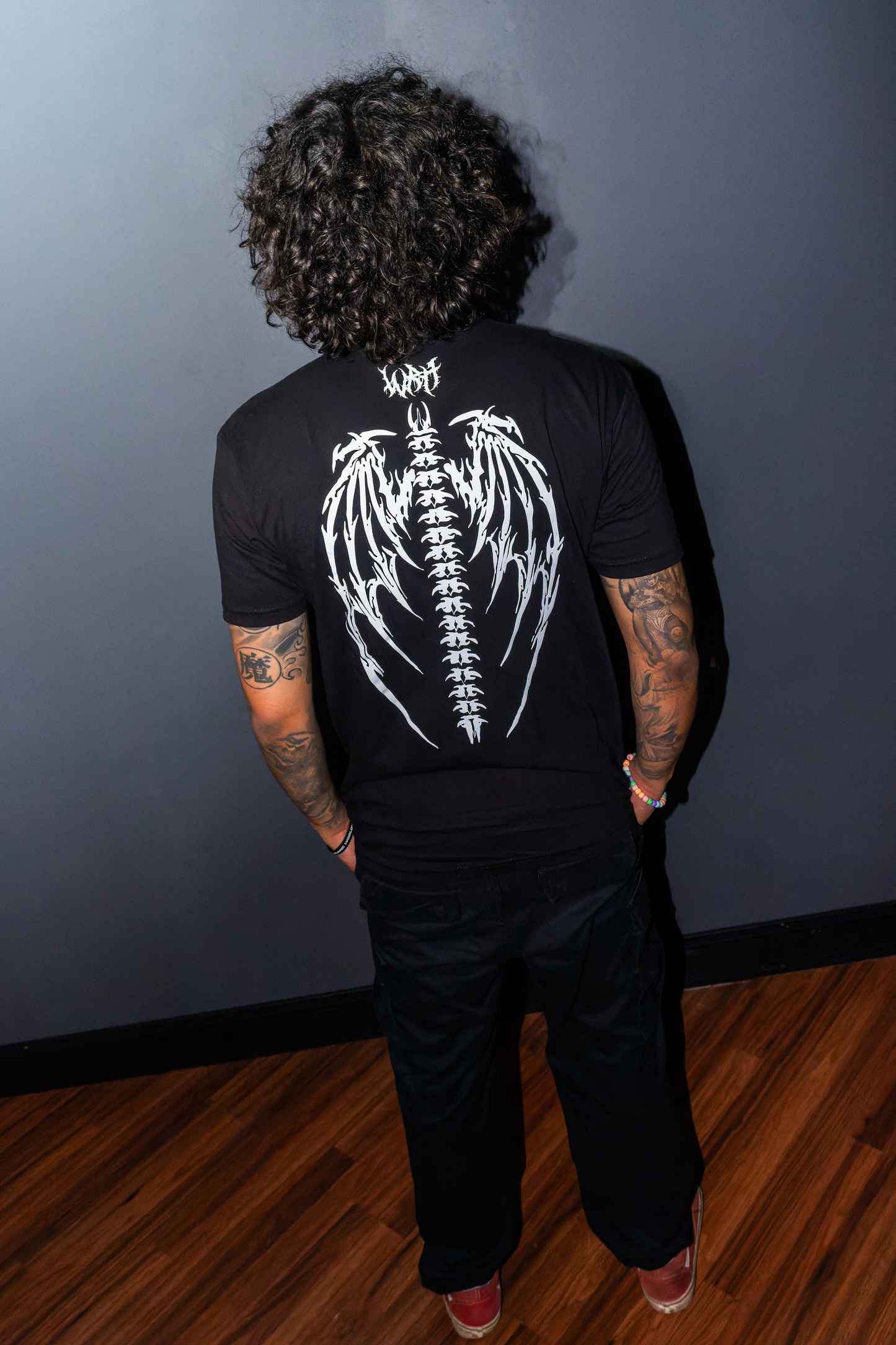 A man viewed from behind wearing the Riddim Fallen Angel Tee t-shirt in black with white winged skeleton graphic on the back, featuring "WRH" Text in Barbed font paired with black pants, standing against a dark gray wall on a wooden floor.