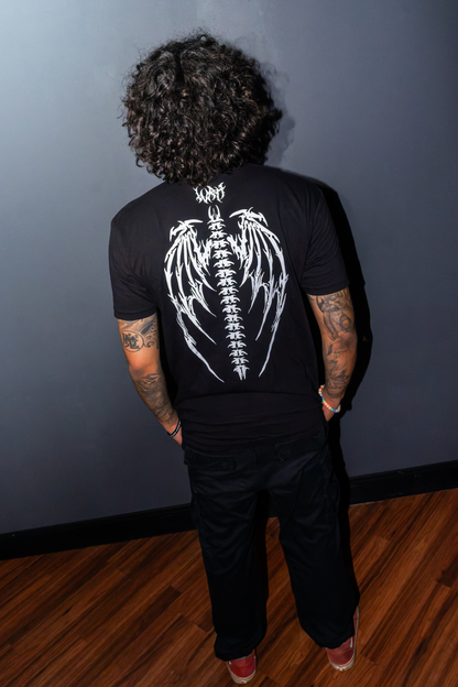 A man viewed from behind wearing the Riddim Fallen Angel Tee t-shirt in black with white winged skeleton graphic on the back, featuring "WRH" Text in Barbed font paired with black pants, standing against a dark gray wall on a wooden floor.