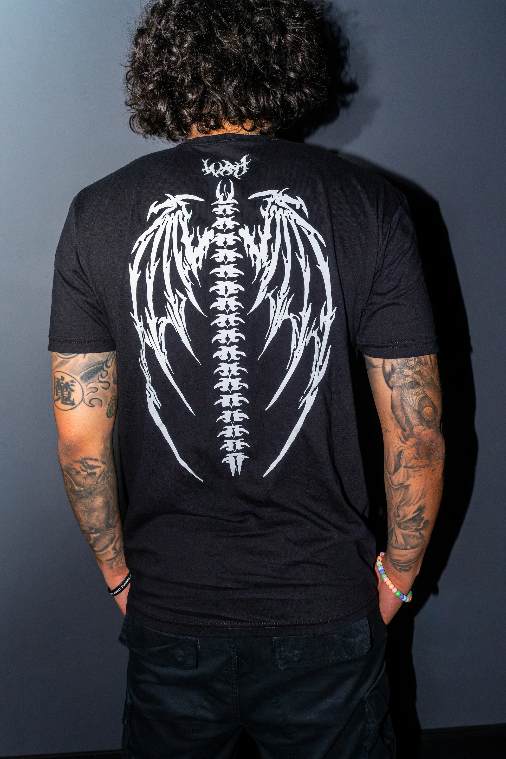 A man viewed from behind wearing the Riddim Fallen Angel t-shirt with a large white winged spine graphic on the back, and "WRH" printed in barbed text above it set against a dark gray background.