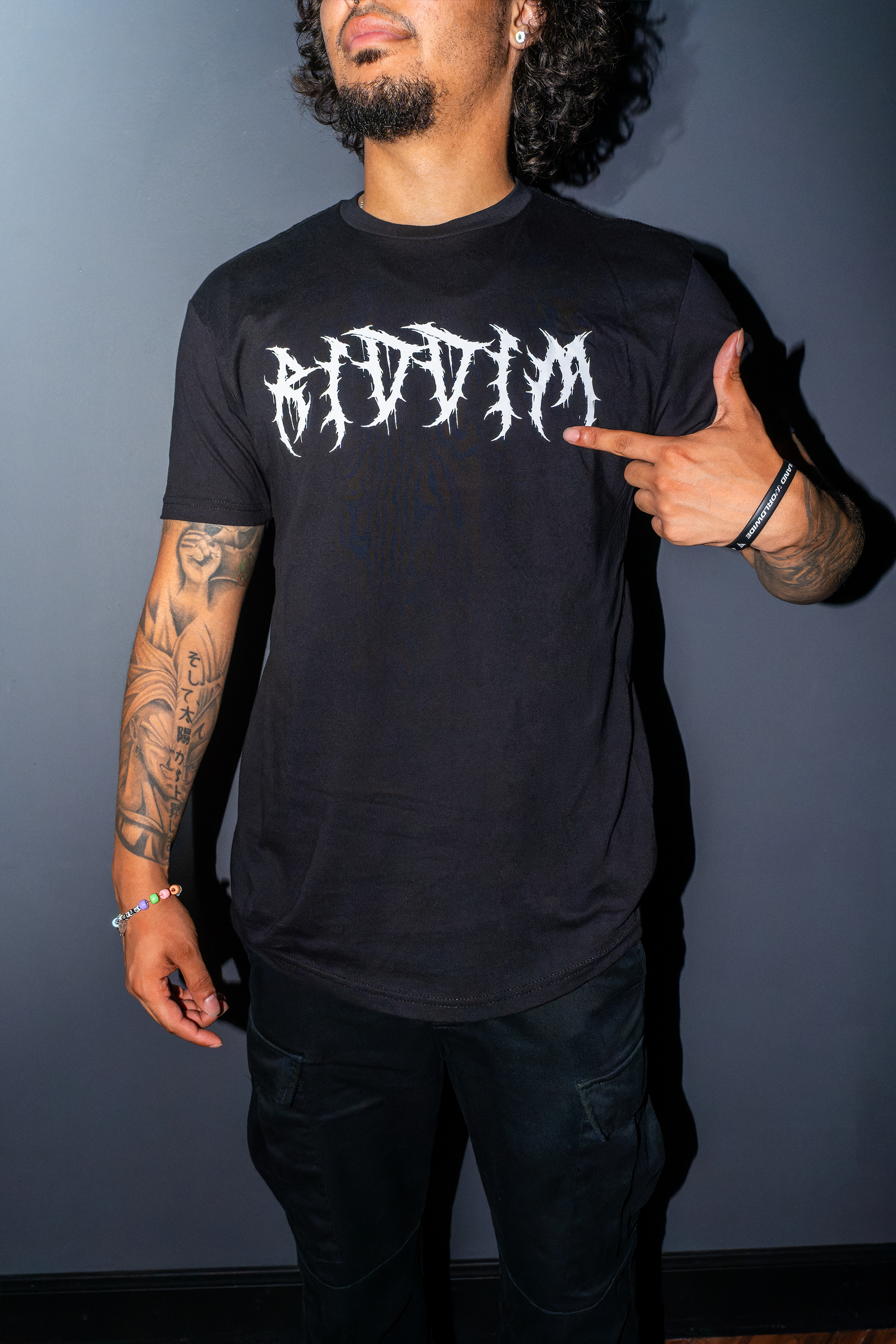 A man wearing the riddim fallen angel  t-shirt with a white barbed gothic-style 'RIDDIM' graphic on the chest, pointing to the text. The background is a solid dark gray wall.