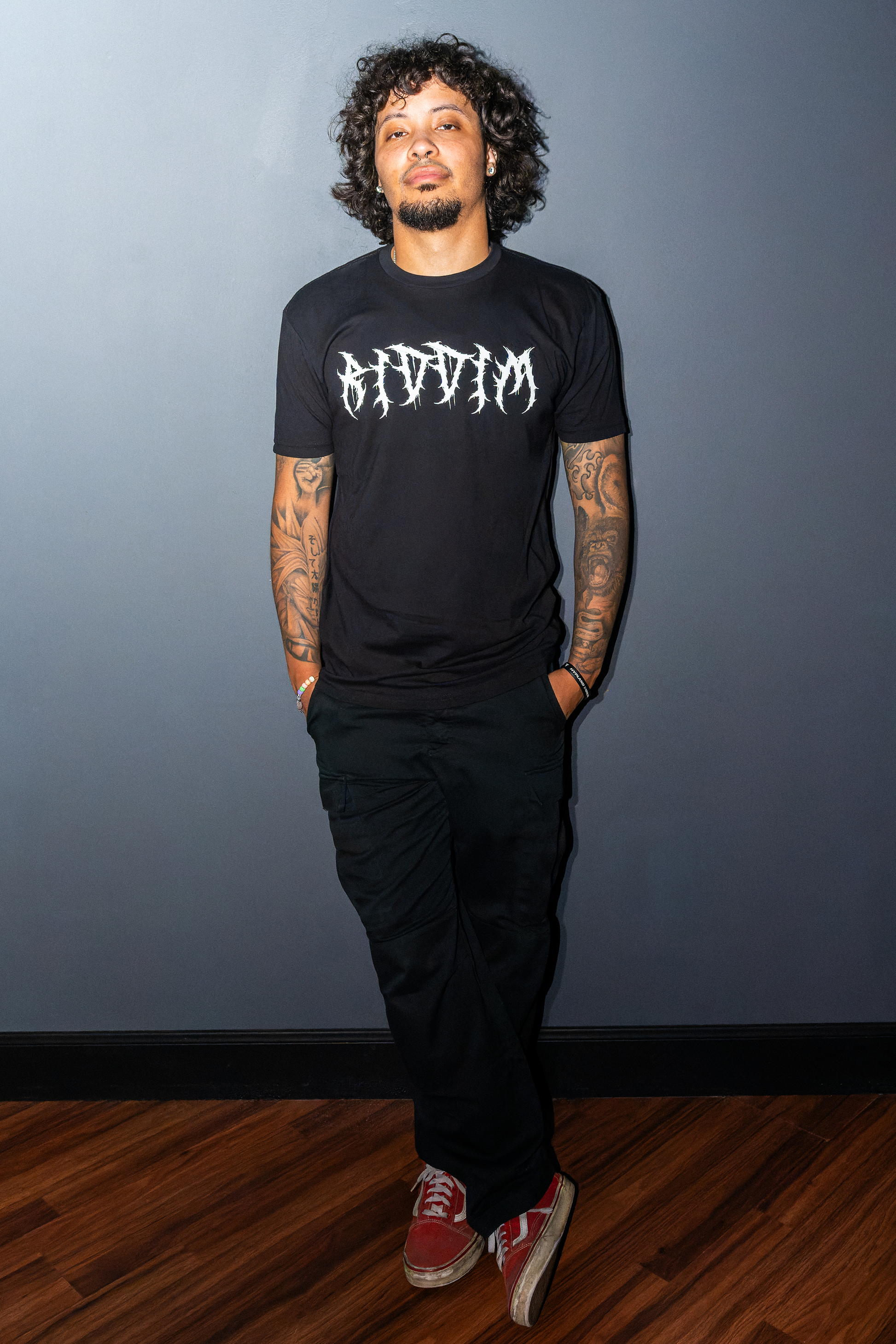 A man with curly hair and tattoos stands against a dark gray wall wearing a black Riddim Fallen Angel with the word 'RIDDIM' in a barbed metal-style white font, black pants, and red sneakers.