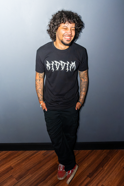 A man with curly dark hair and tattoos stands smiling, wearing a black graphic Riddim Fallen Angel t-shirt with the word 'RIDDIM' in a stylized white font, black pants, and red sneakers, posing against a dark gray wall and wood floor.