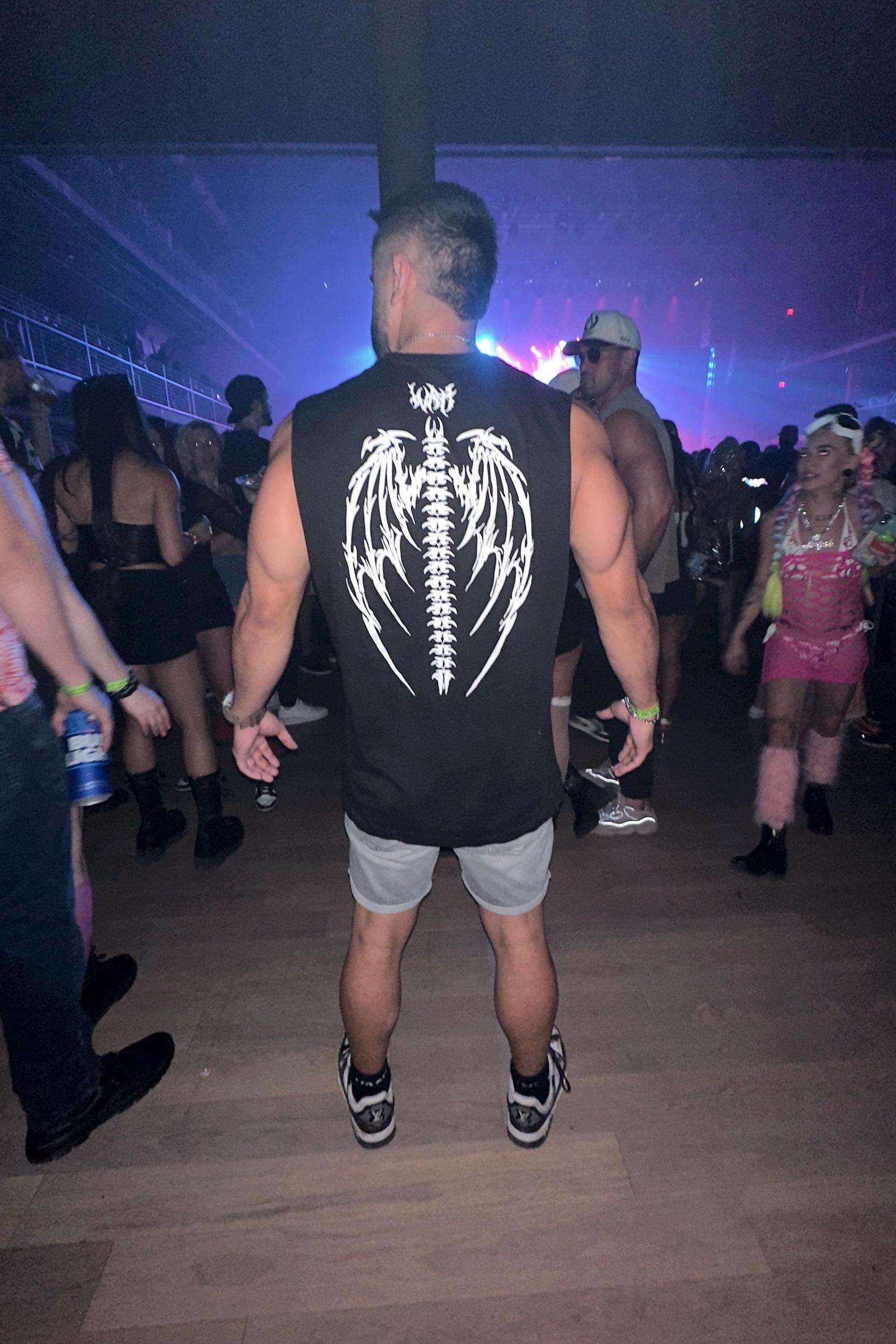 A person wearing a black sleeveless t-shirt with a graphic of a fallen angel on the back, standing in a crowd at a rave.