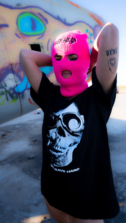 A person wearing a bright pink knit balaclava mask with "BASS HEAD" printed on the forehead, and a black graphic t-shirt with a white skull print with text "WE RAVE HARD," posing outdoors in front of a graffiti-covered wall.